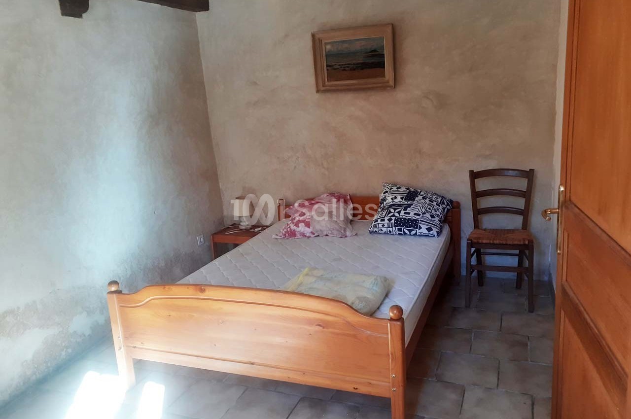 Chambre simple avec un lit en bois, une chaise, un tableau au mur et un sol en carrelage clair.