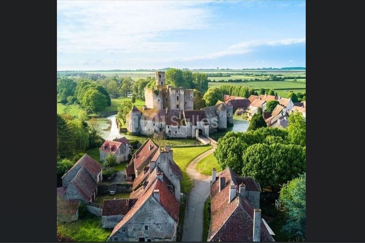 Vue aérienne d'un village avec un château médiéval entouré d'eau, des maisons aux toits rouges et des champs verdoyants.