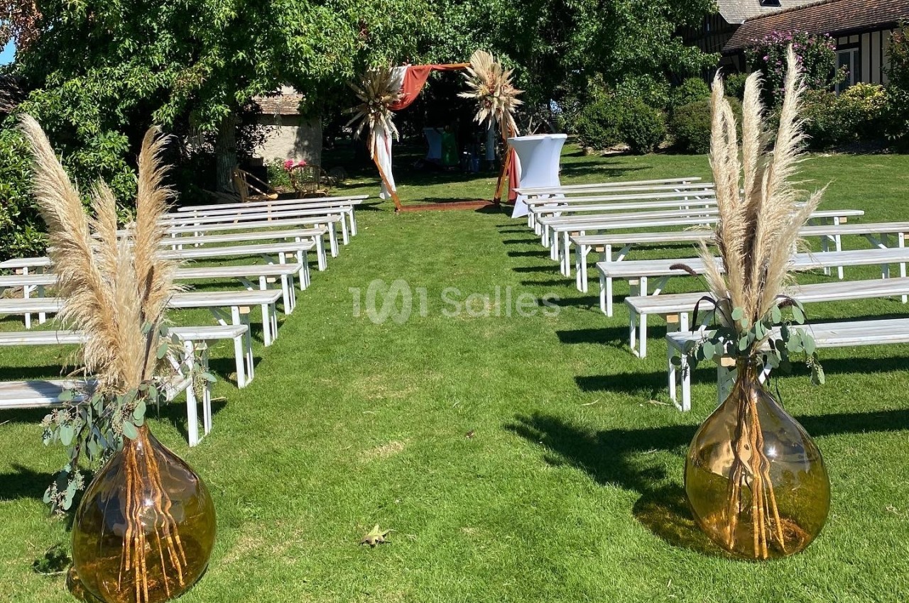 Allée de cérémonie extérieure avec bancs blancs, décorée de vases ambrés et de pampas, sur une pelouse verdoyante.