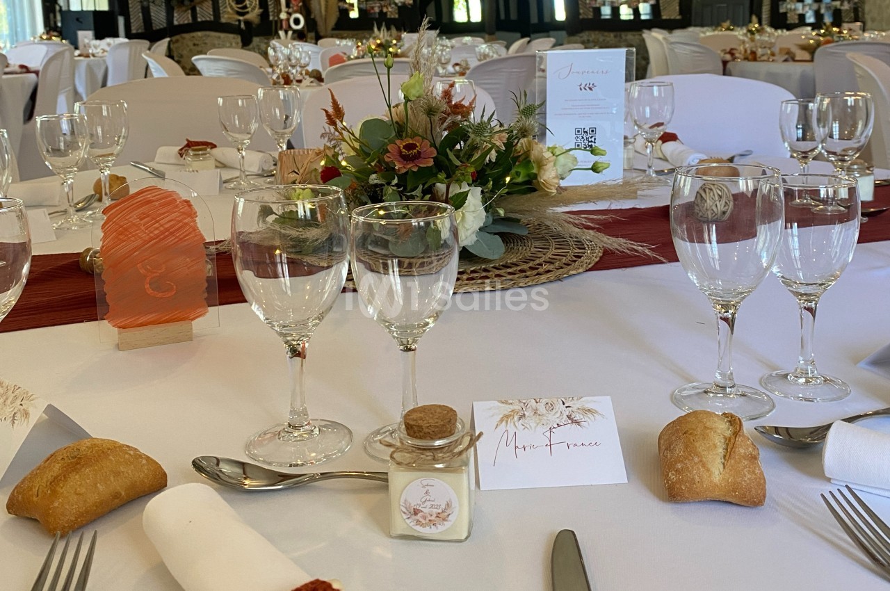 Table de réception décorée avec des fleurs, menus, verres, couverts et petits pains disposés sur une nappe blanche.