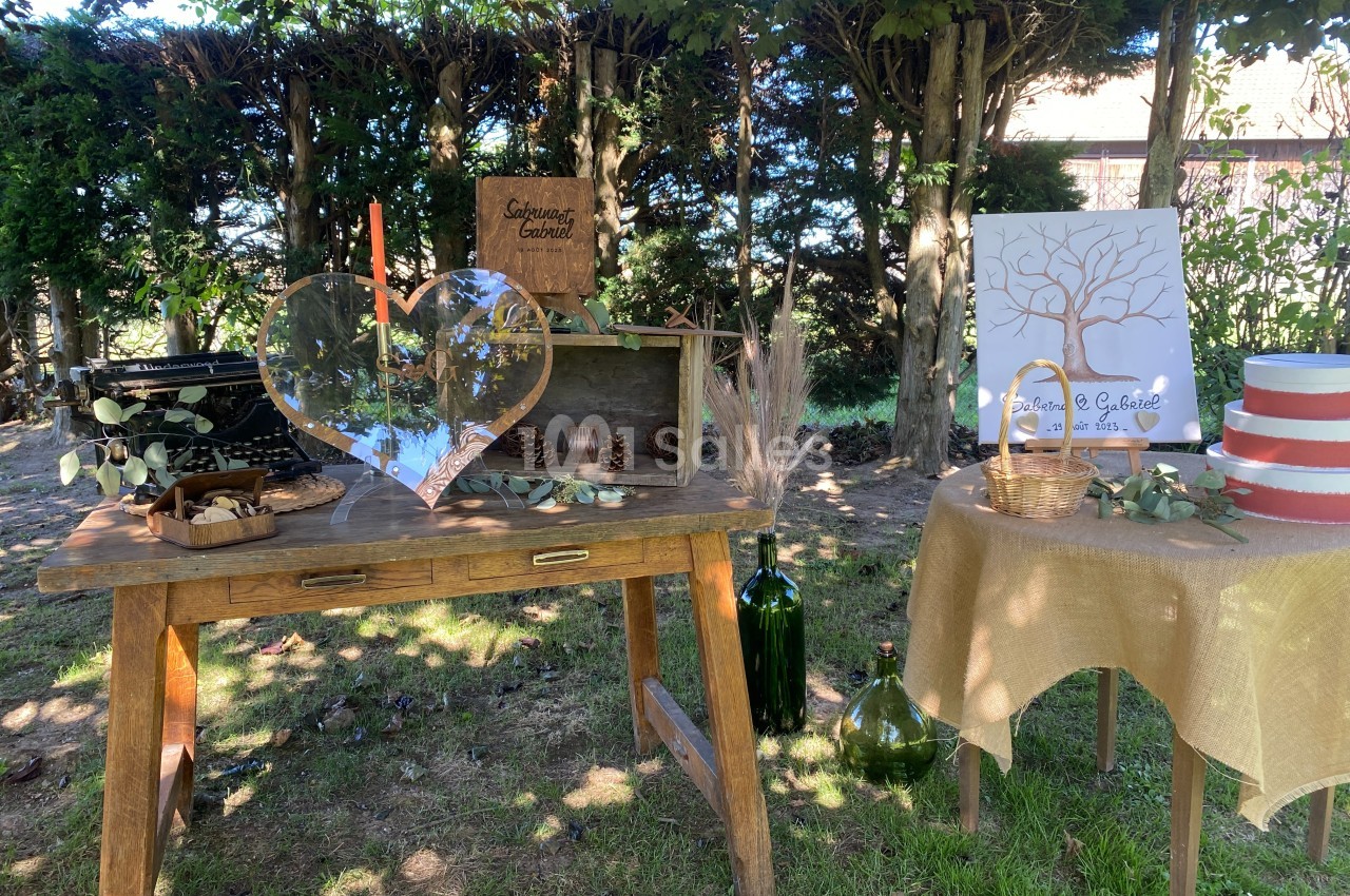 Décoration champêtre avec table en bois, objets rustiques, cœur en verre et arbre à empreintes sous des arbres.