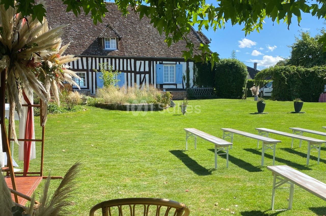 Jardin verdoyant avec bancs blancs disposés devant une maison à colombages sous un ciel bleu.