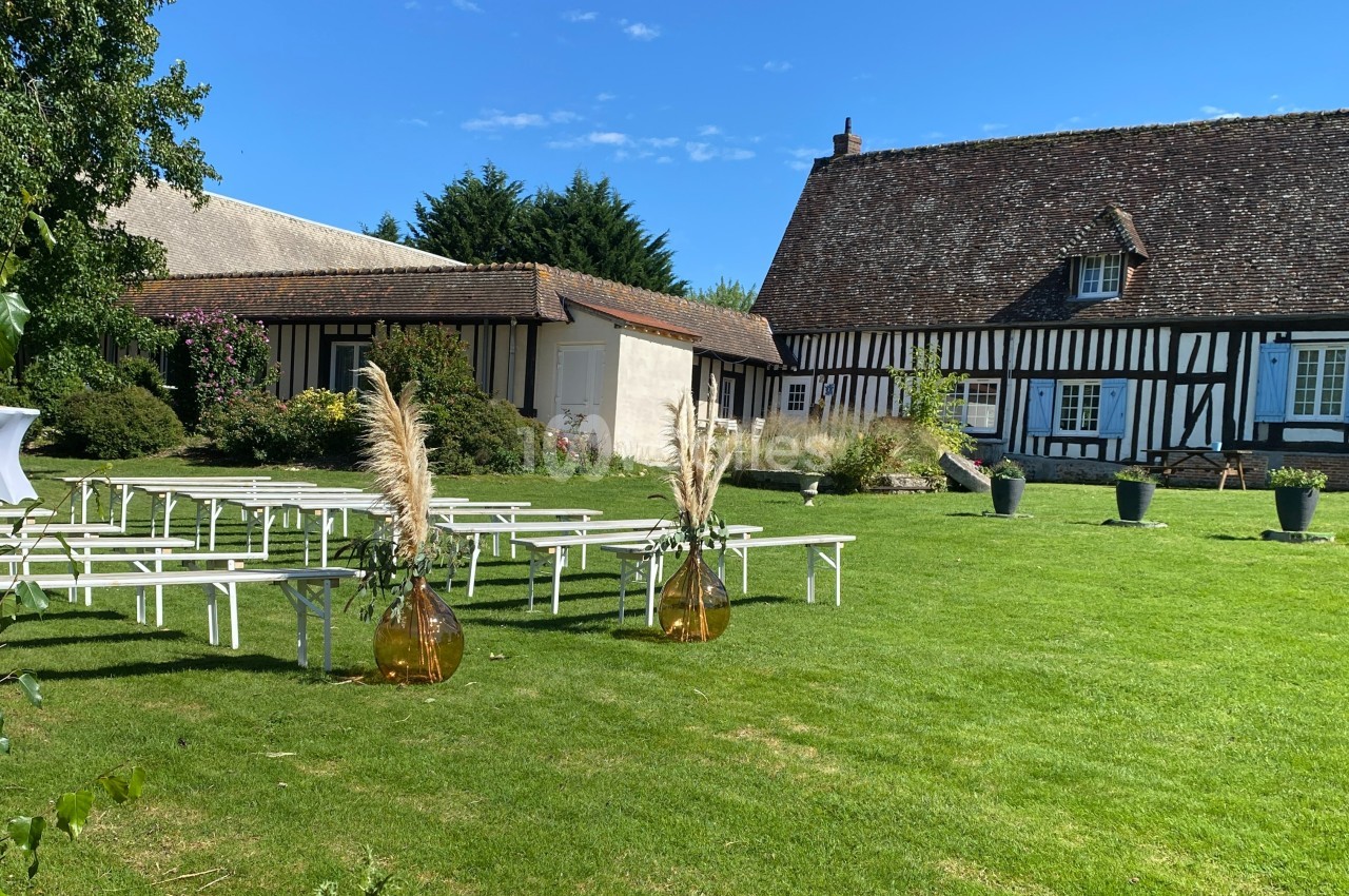 Jardin avec pelouse aménagée pour un événement, bancs blancs alignés et décorations florales devant un bâtiment à colombages.