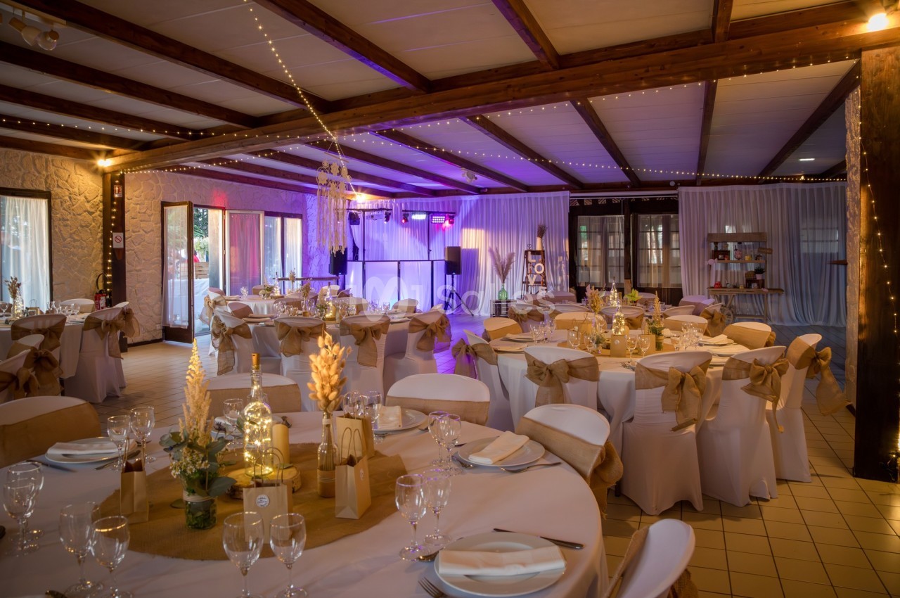 Salle de réception décorée avec des tables rondes, nappes blanches, chaises ornées de nœuds beige et éclairage chaleureux.
