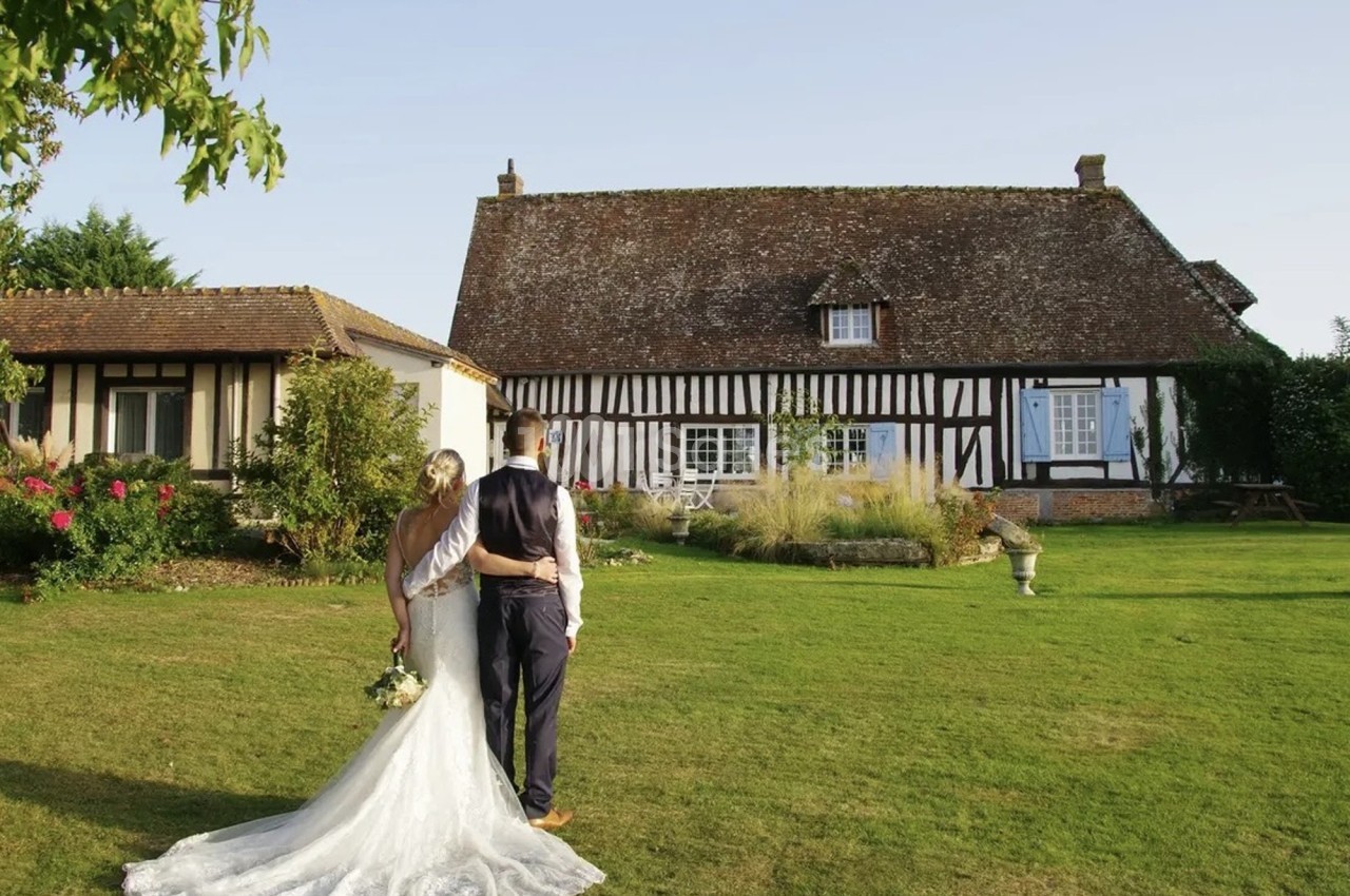 Un couple en tenue de mariage regarde une maison à colombages dans un jardin verdoyant par une journée ensoleillée.