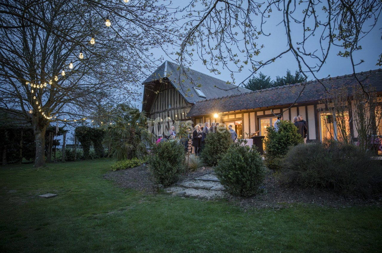 Maison à colombages éclairée en soirée, entourée d'arbres et de pelouse, avec des invités rassemblés à l'extérieur.