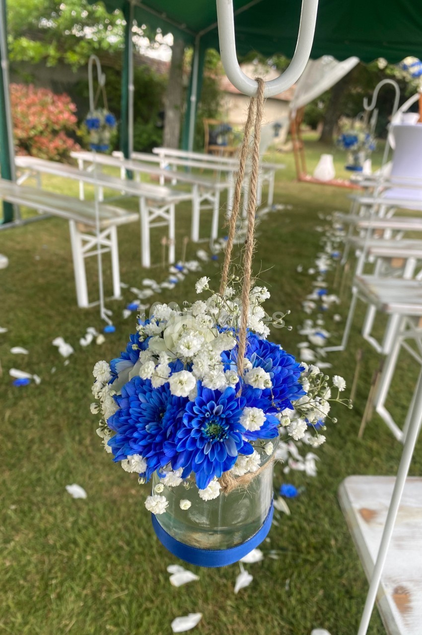 Bouquet de fleurs bleues et blanches suspendu dans un bocal en verre, décorant une allée extérieure pour un événement.