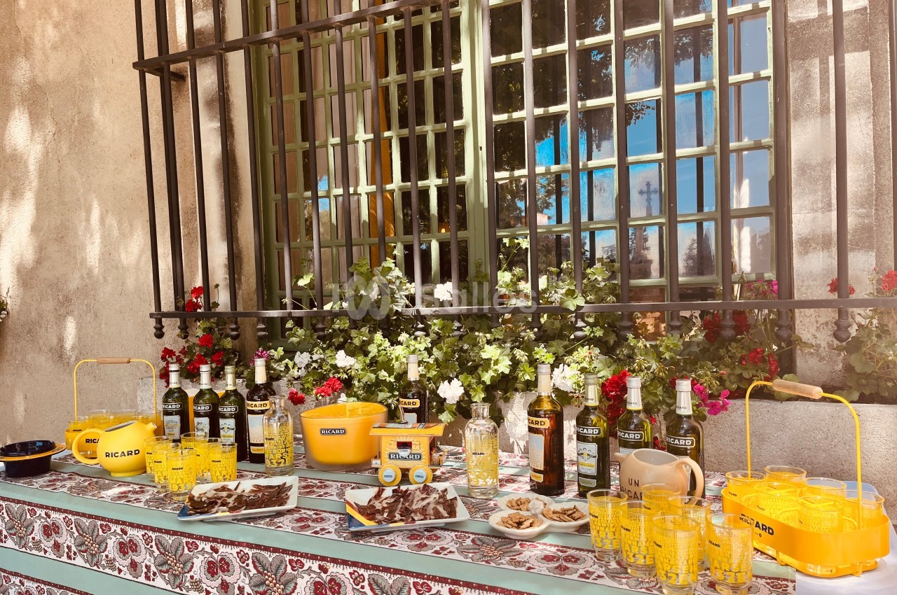 Table dressée en extérieur avec bouteilles, verres, apéritifs et décorations florales devant une fenêtre grillagée.