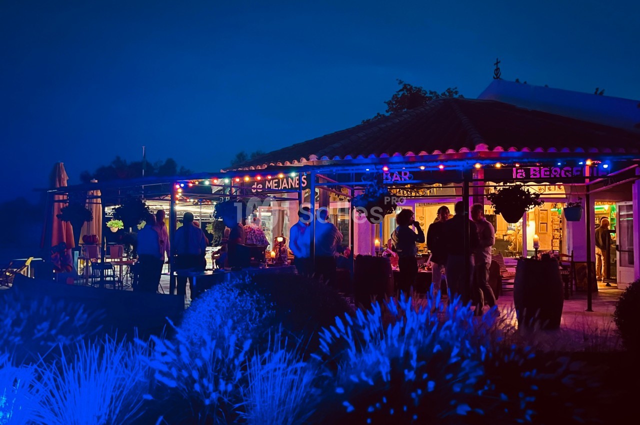 Terrasse d'un bar-restaurant éclairée par des lumières colorées en soirée, avec des clients debout et assis.