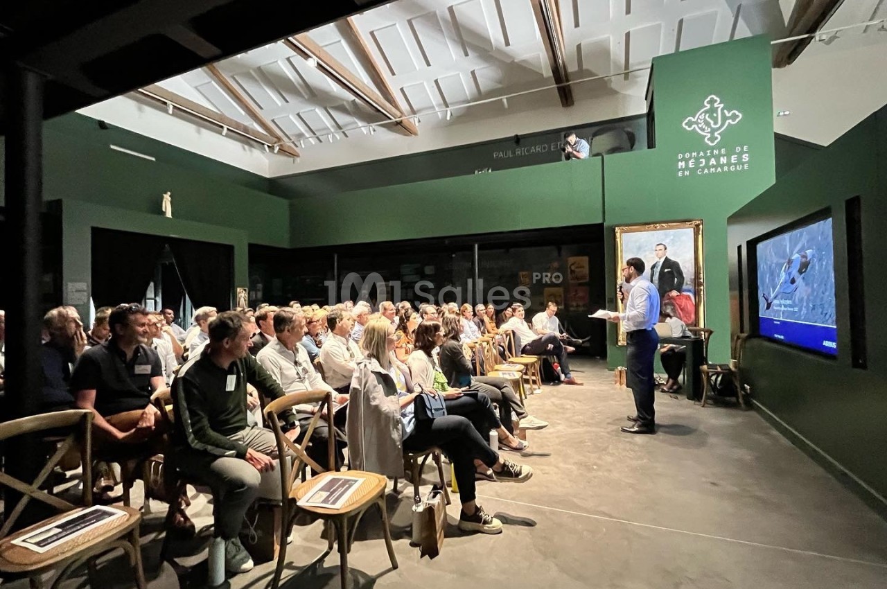 Un conférencier s'adresse à un public assis dans une salle sombre avec un écran et un tableau en arrière-plan.