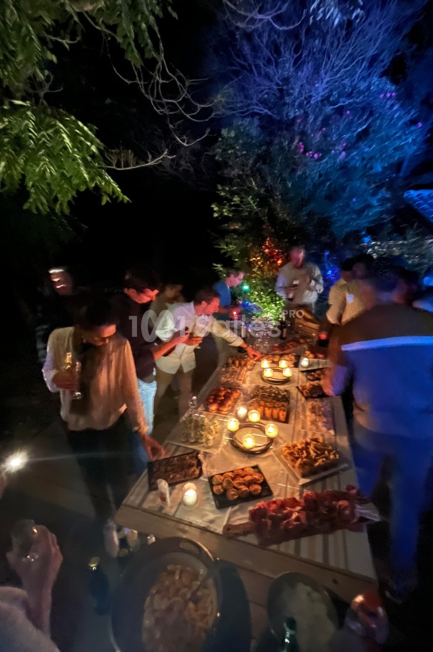 Des personnes se servent autour d'une grande table garnie de plats variés, éclairée par des bougies, en extérieur de nuit.