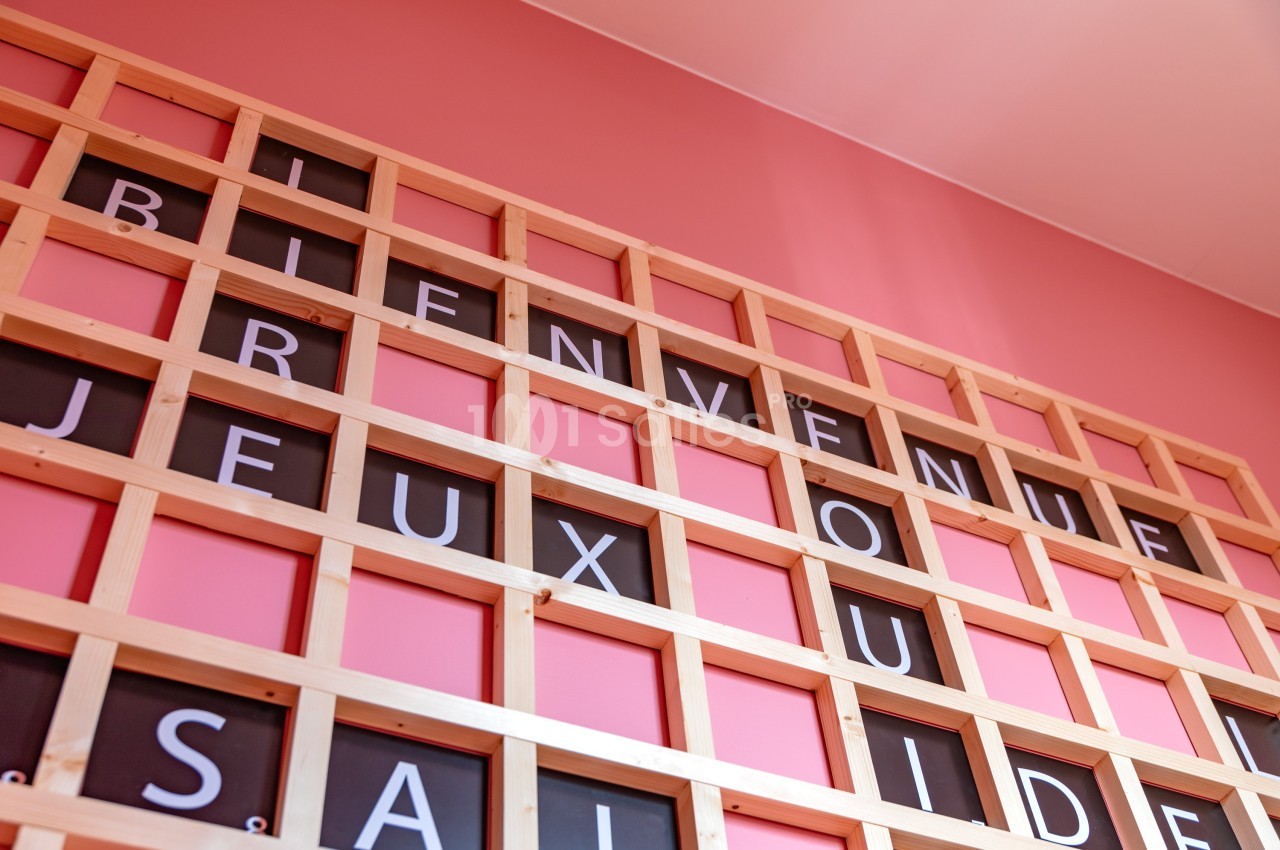 Mur rose avec une grille en bois affichant des lettres formant des mots comme ’Bienvenue’ et ’Jeux’.