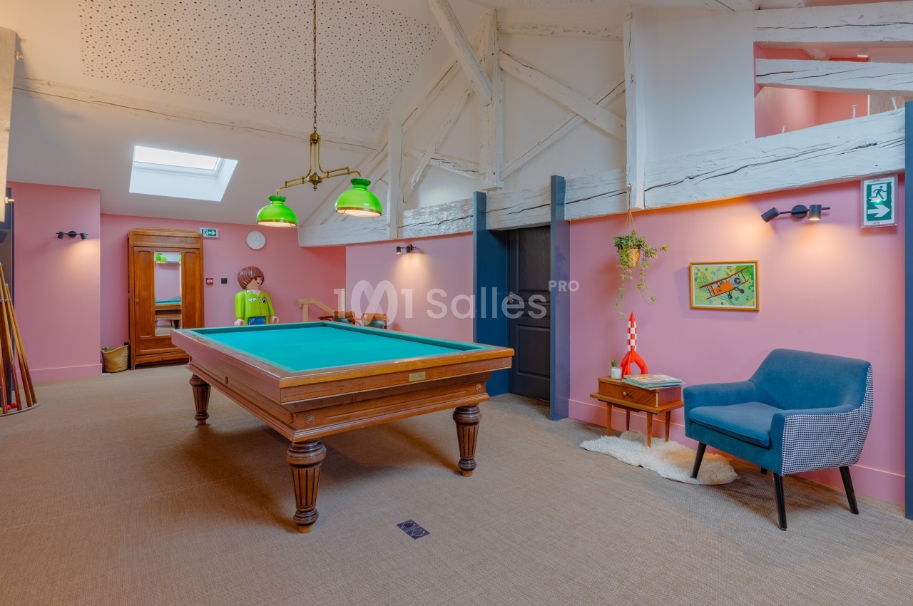 Salle de jeux avec une table de billard, fauteuil bleu, décoration murale et poutres apparentes sous un plafond mansardé.