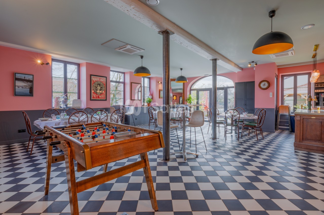 Salle lumineuse avec baby-foot, tables et chaises, sol en damier noir et blanc, murs roses et grandes fenêtres.