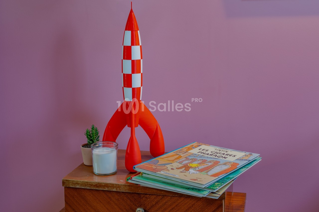 Maquette de fusée rouge et blanche posée sur une table en bois avec une plante, une bougie et des livres pour enfants.