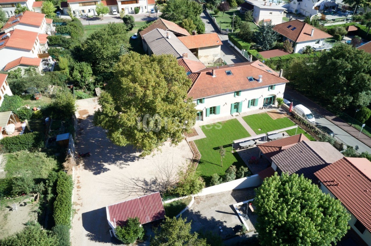 Vue aérienne d'une maison avec jardin, allée gravillonnée, arbres et bâtiments voisins dans un quartier résidentiel.