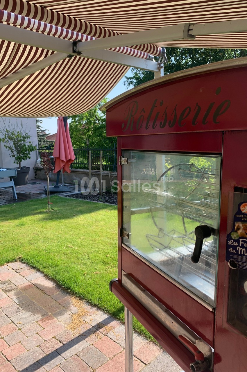 Rôtissoire rouge sous un auvent rayé, installée dans un jardin avec pelouse, terrasse et mobilier extérieur.