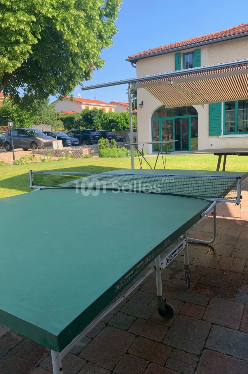 Table de ping-pong verte installée en extérieur sur une terrasse pavée, avec une maison et un jardin en arrière-plan.