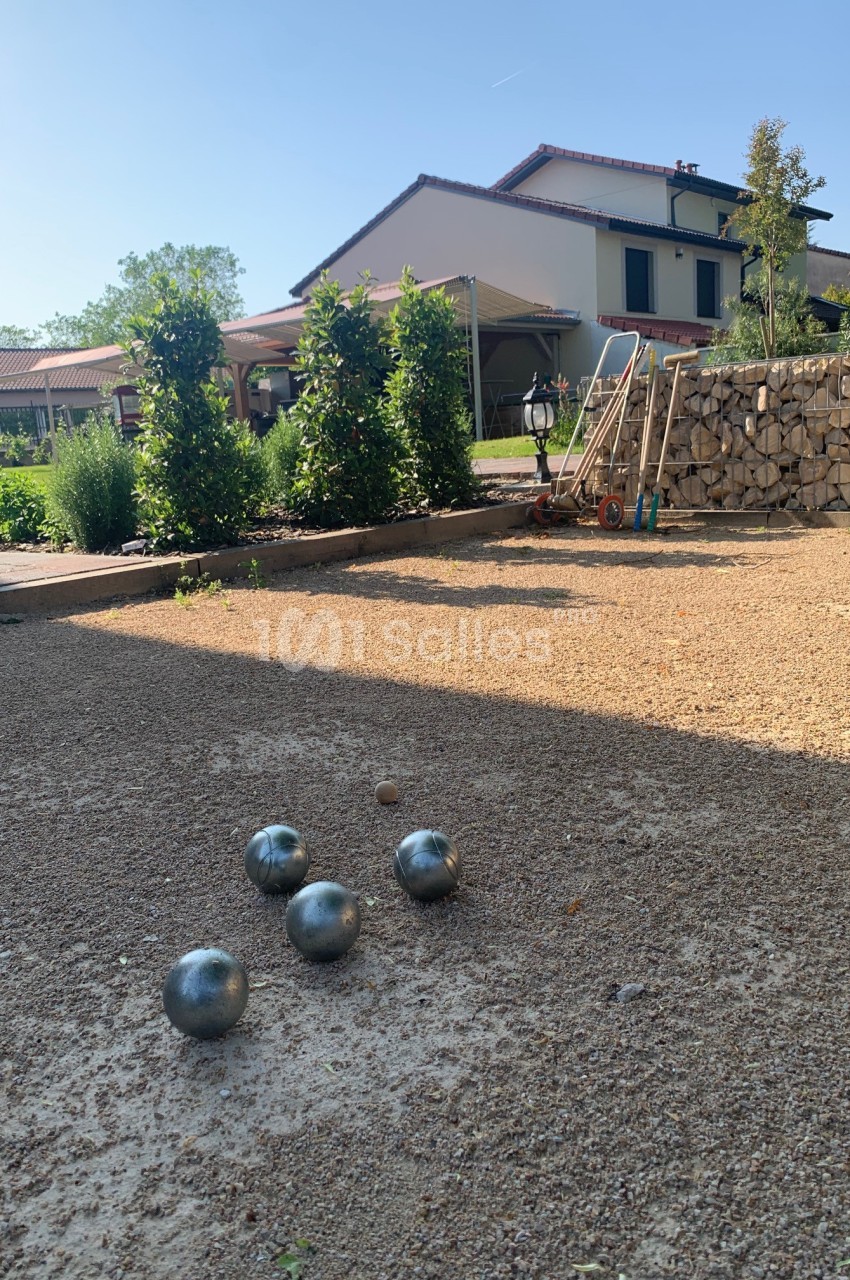 Terrain de pétanque avec des boules en métal et un cochonnet, entouré de végétation et d'une maison en arrière-plan.
