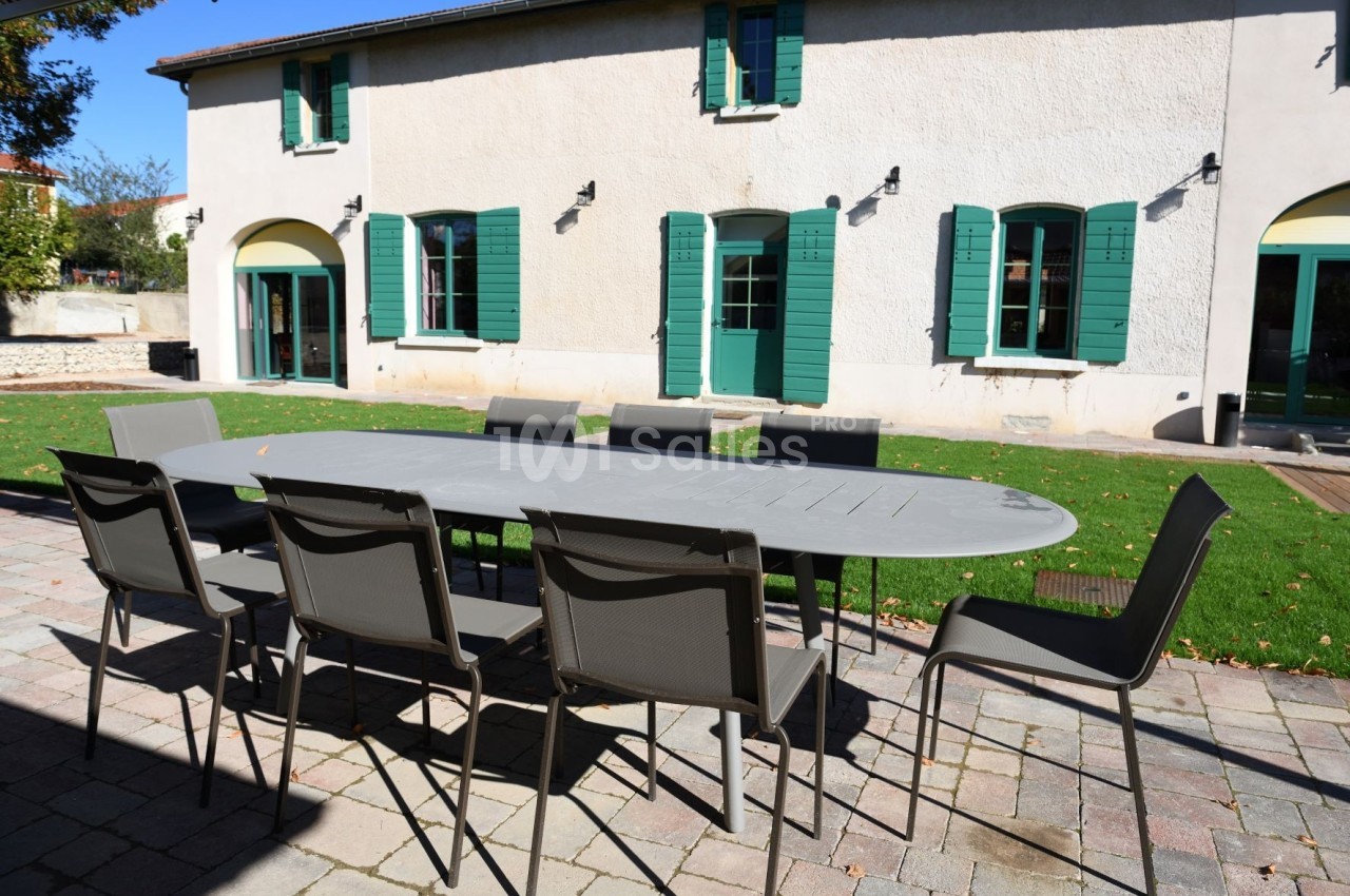 Table de jardin grise entourée de chaises noires sur une terrasse en pavés, devant une maison aux volets verts.