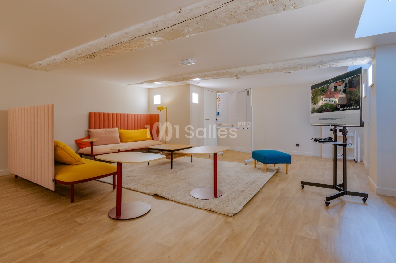 Salle lumineuse avec parquet, mobilier coloré, poutres apparentes et écran sur pied dans un espace aménagé.