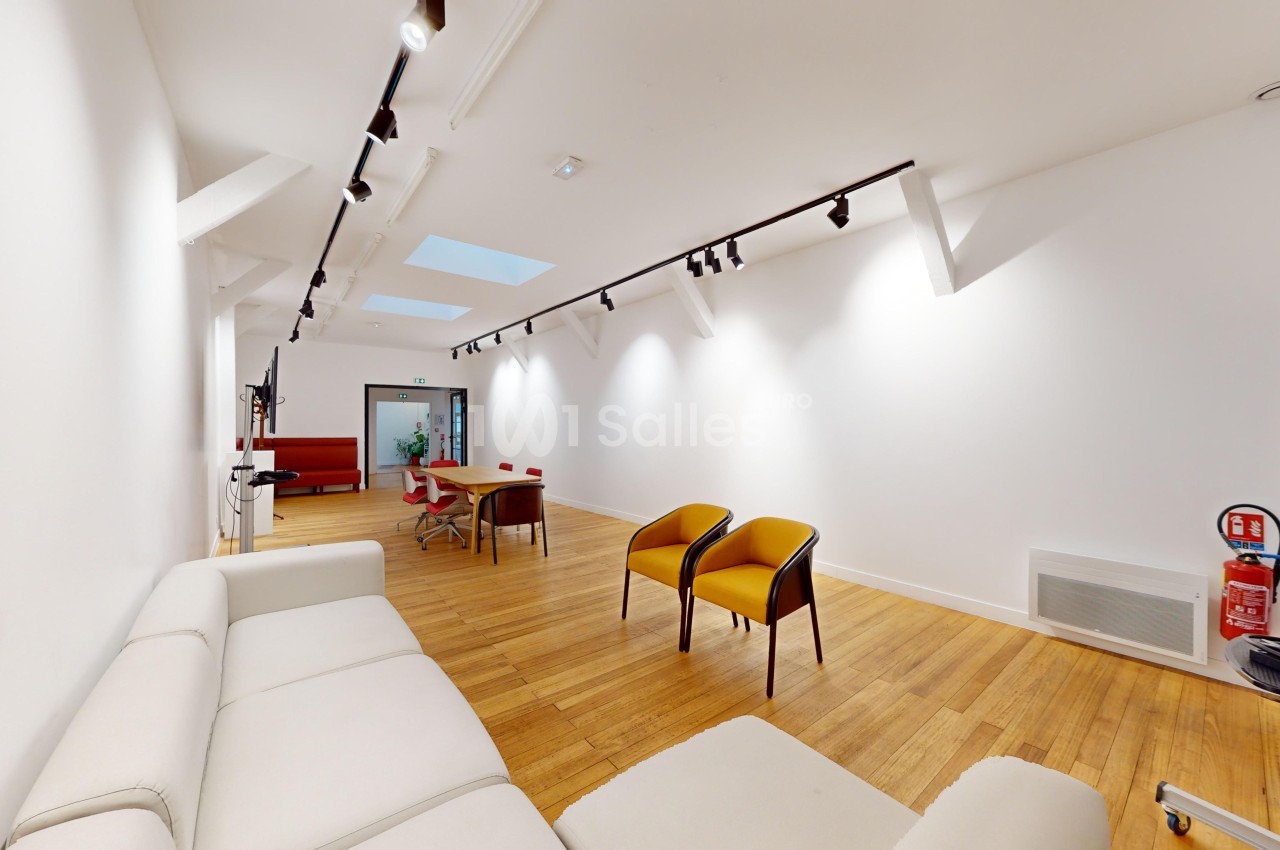 Salle lumineuse avec parquet, mobilier moderne, canapés blancs, chaises colorées et éclairage au plafond.