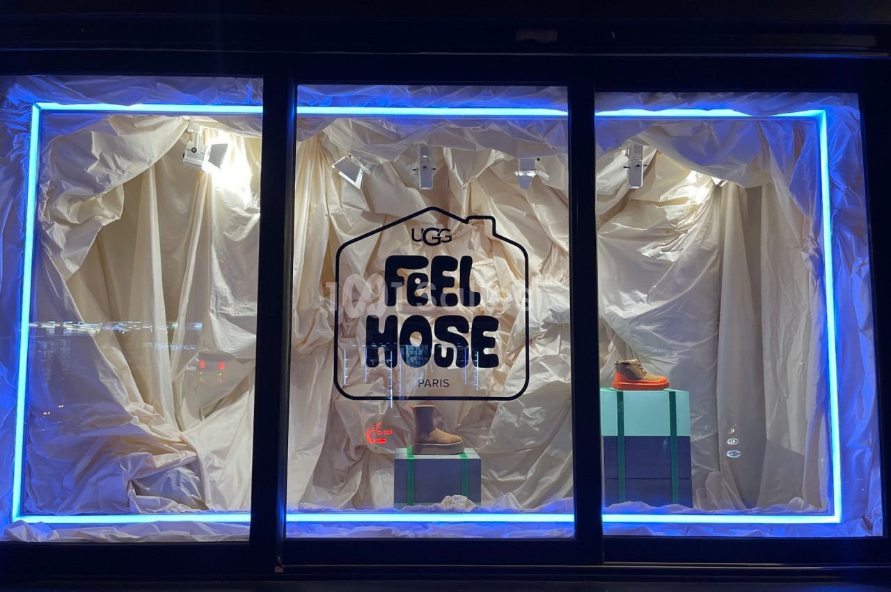 Vitrine éclairée avec des chaussures exposées sur des socles, entourées de rideaux beige et encadrée de néons bleus.