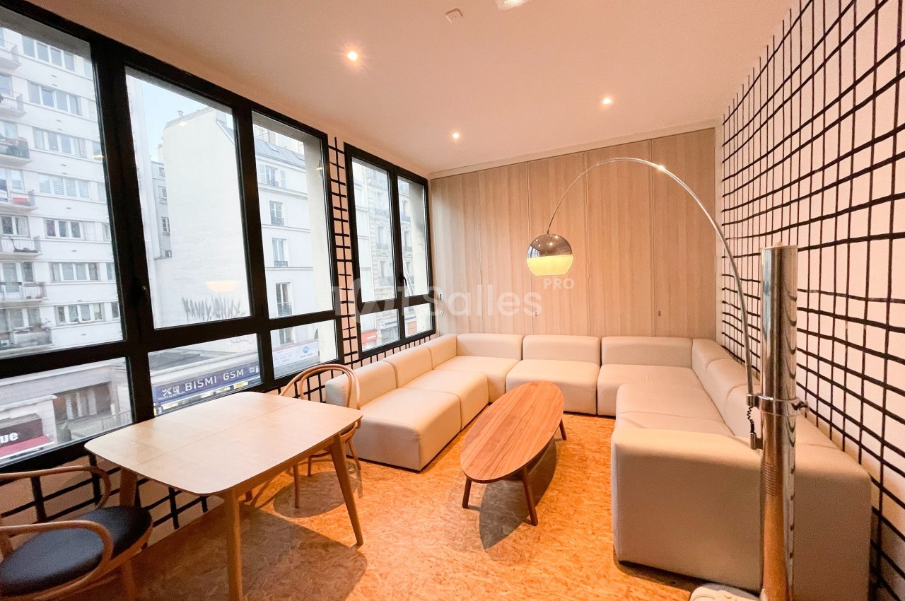 Salle lumineuse avec canapé d'angle beige, table basse en bois, table à manger et grandes fenêtres donnant sur la ville.