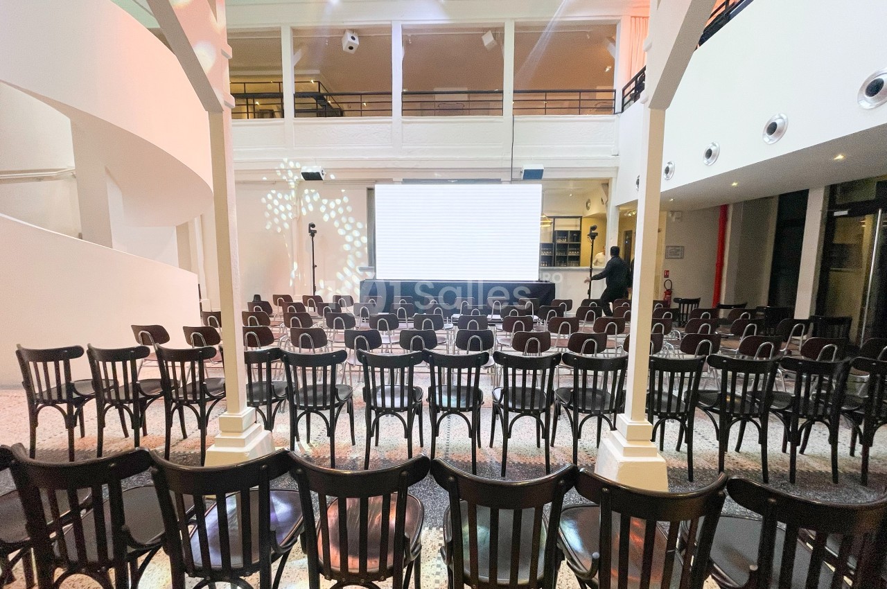 Salle de conférence avec des rangées de chaises noires face à un écran blanc, éclairage tamisé.