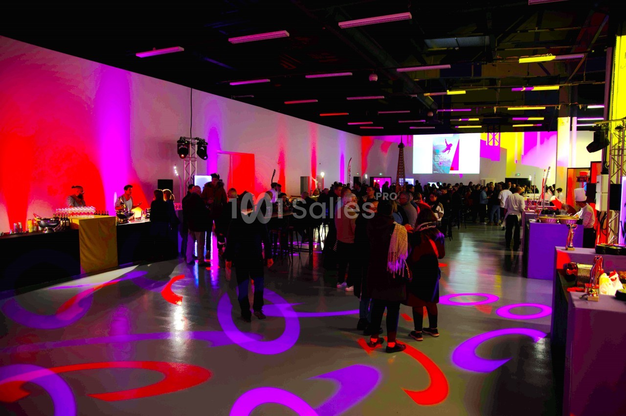 Salle événementielle avec éclairage coloré, tables hautes, buffet et invités discutant dans une ambiance festive.