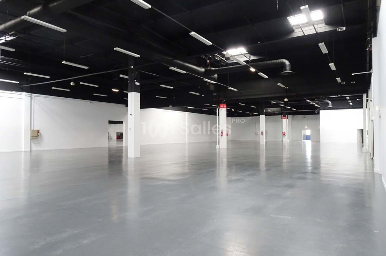 Salle vide avec sol en béton gris, murs blancs et plafond noir équipé de luminaires suspendus.