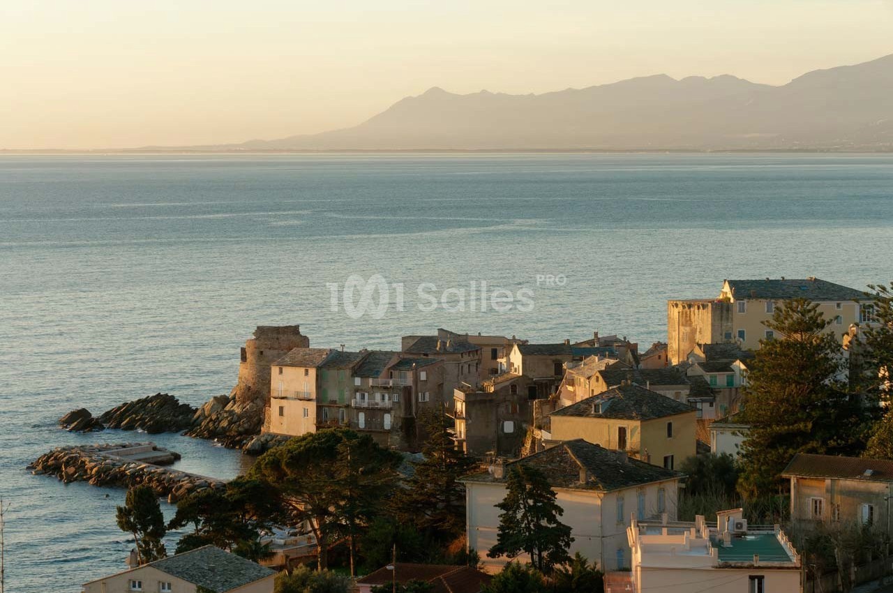 Village côtier avec maisons en pierre, bordé par la mer calme au coucher du soleil, montagnes en arrière-plan.