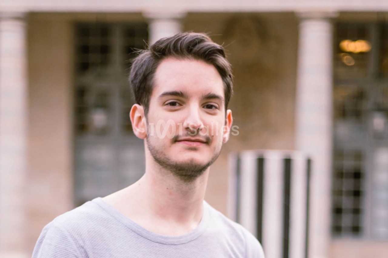 Un homme debout devant un bâtiment avec des colonnes et des fenêtres, portant un t-shirt clair.