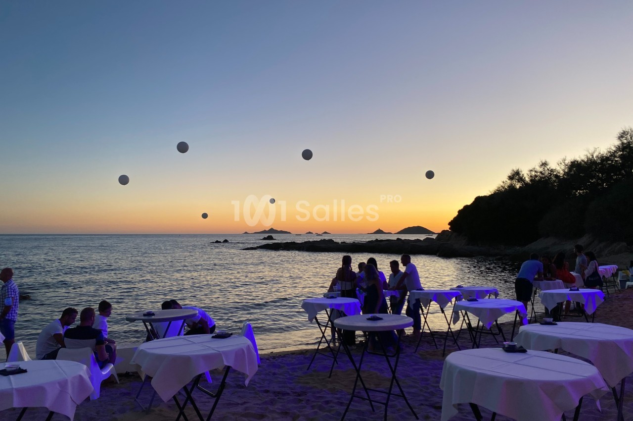 Tables dressées sur une plage au coucher du soleil, avec des invités et des décorations lumineuses flottantes.