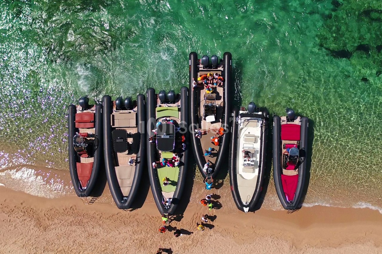 Vue aérienne de plusieurs bateaux pneumatiques alignés sur une plage de sable, avec des personnes à bord et autour.