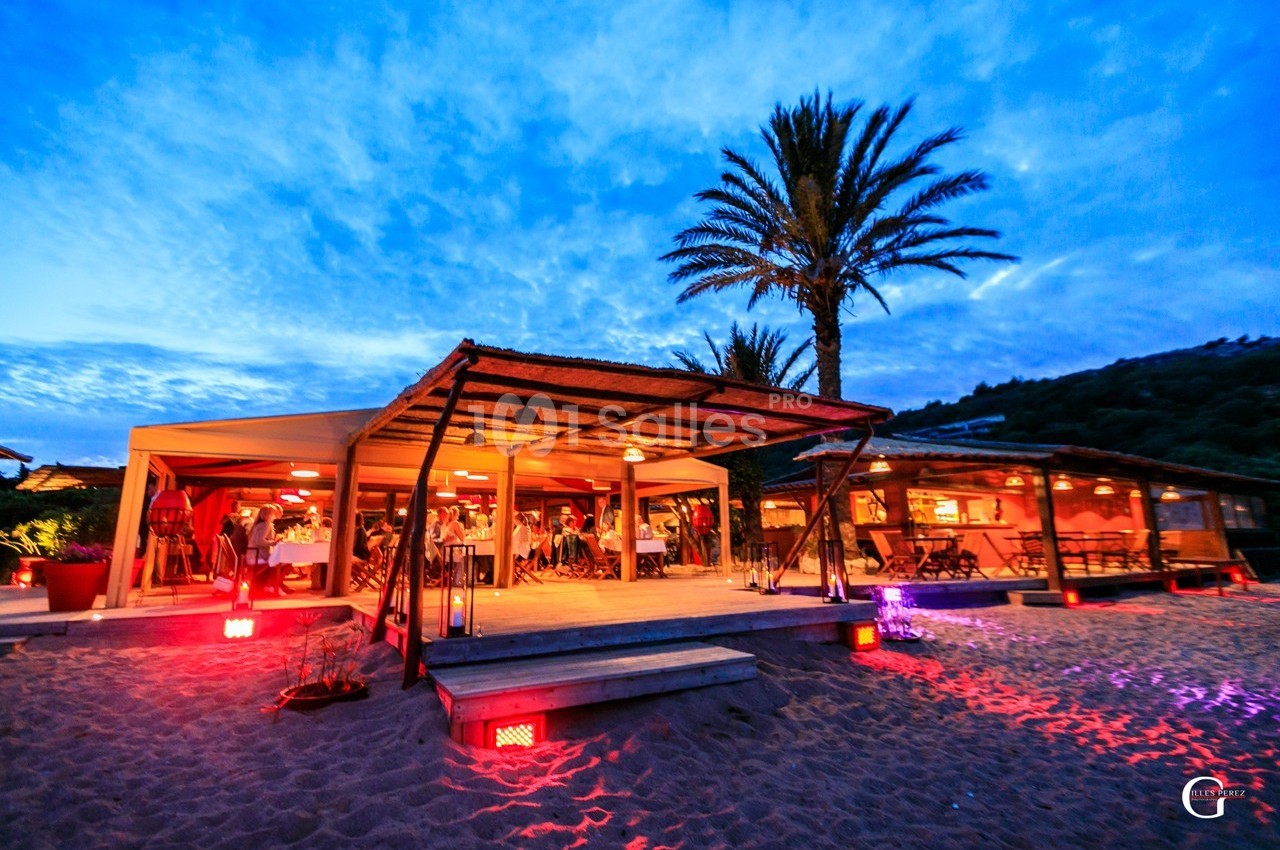 Restaurant en bord de plage avec terrasse éclairée, palmiers et ciel crépusculaire en arrière-plan.