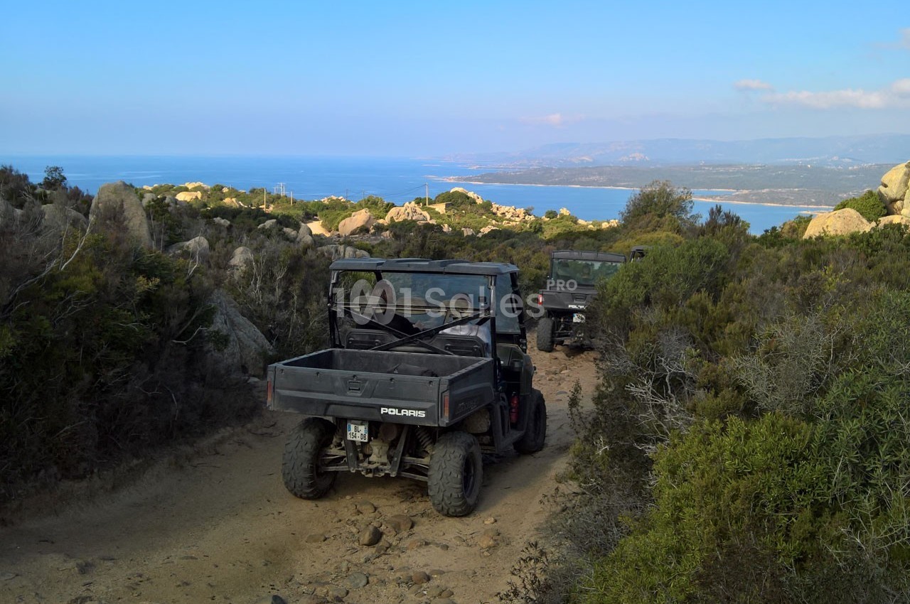 Deux véhicules tout-terrain circulent sur un chemin rocailleux entouré de végétation, avec vue sur la mer au loin.