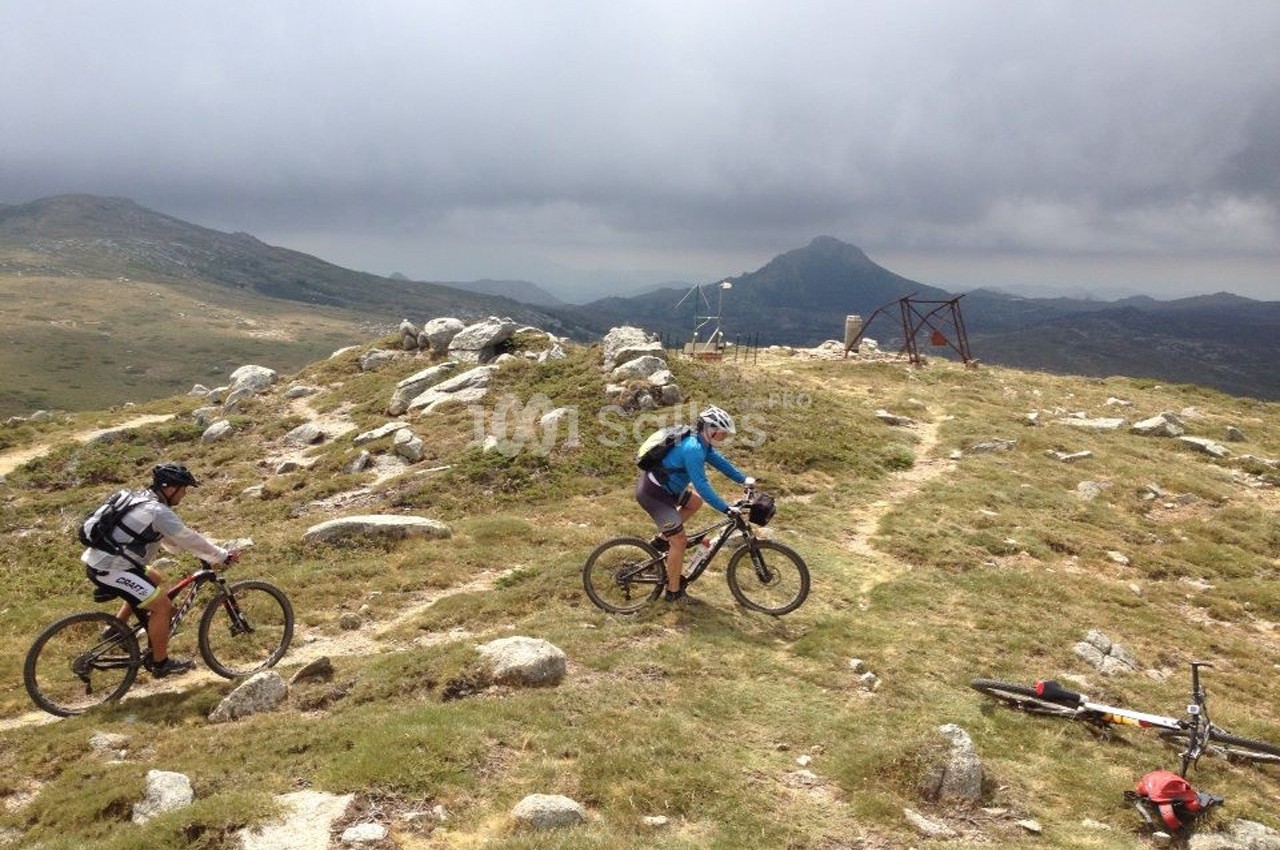 Deux cyclistes en VTT sur un sentier montagneux par temps nuageux, avec un sommet visible à l'horizon.