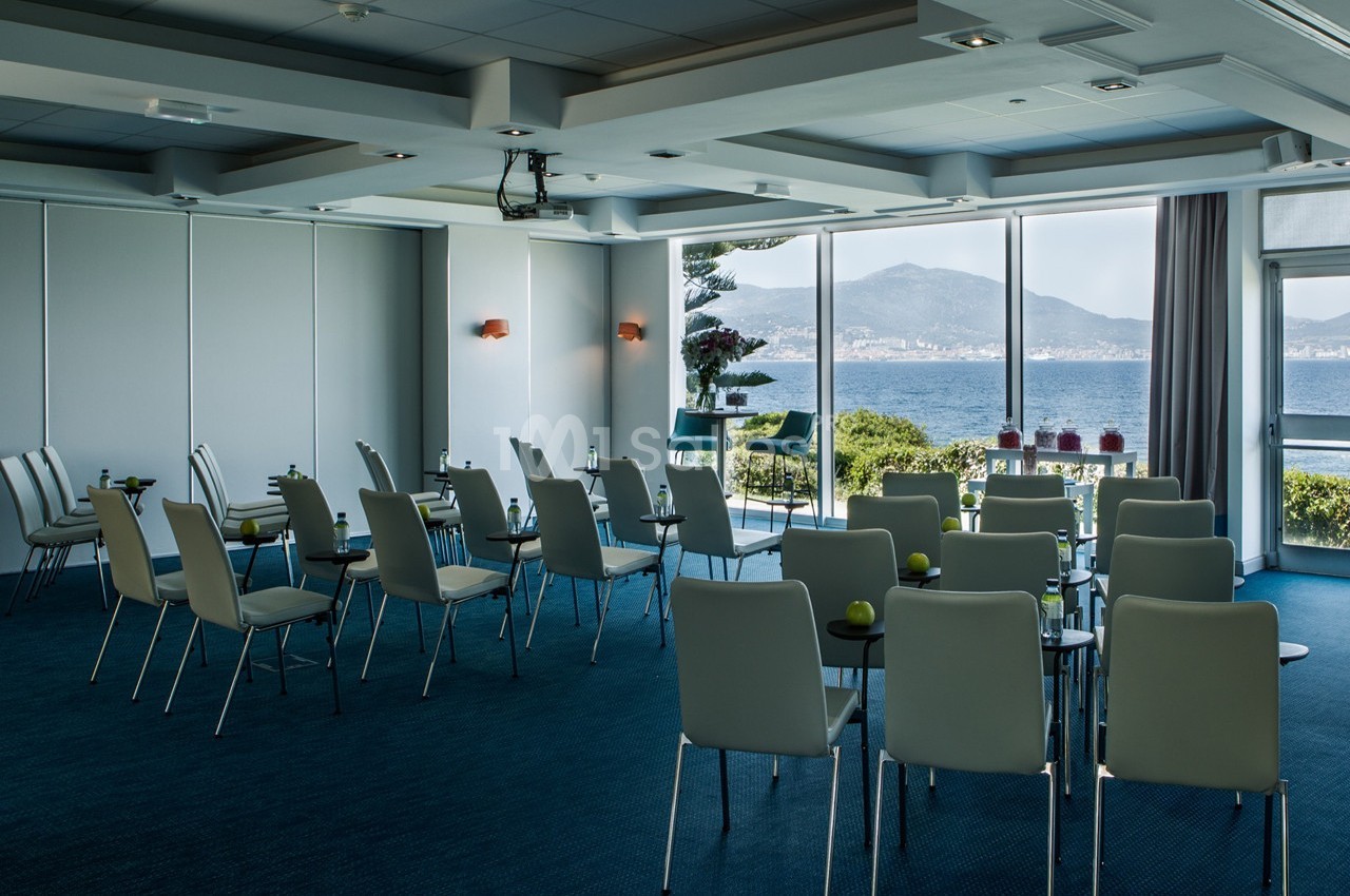 Salle de réunion lumineuse avec chaises disposées en rangées, grandes fenêtres offrant une vue sur la mer et les montagnes.