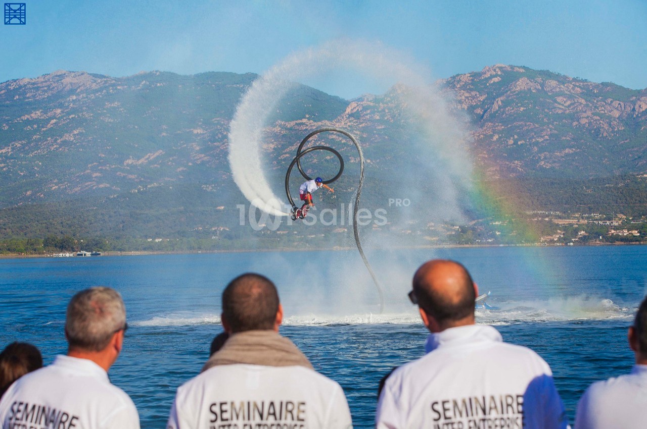 Un homme effectue une figure en flyboard au-dessus de l'eau, sous le regard de spectateurs en chemises blanches.