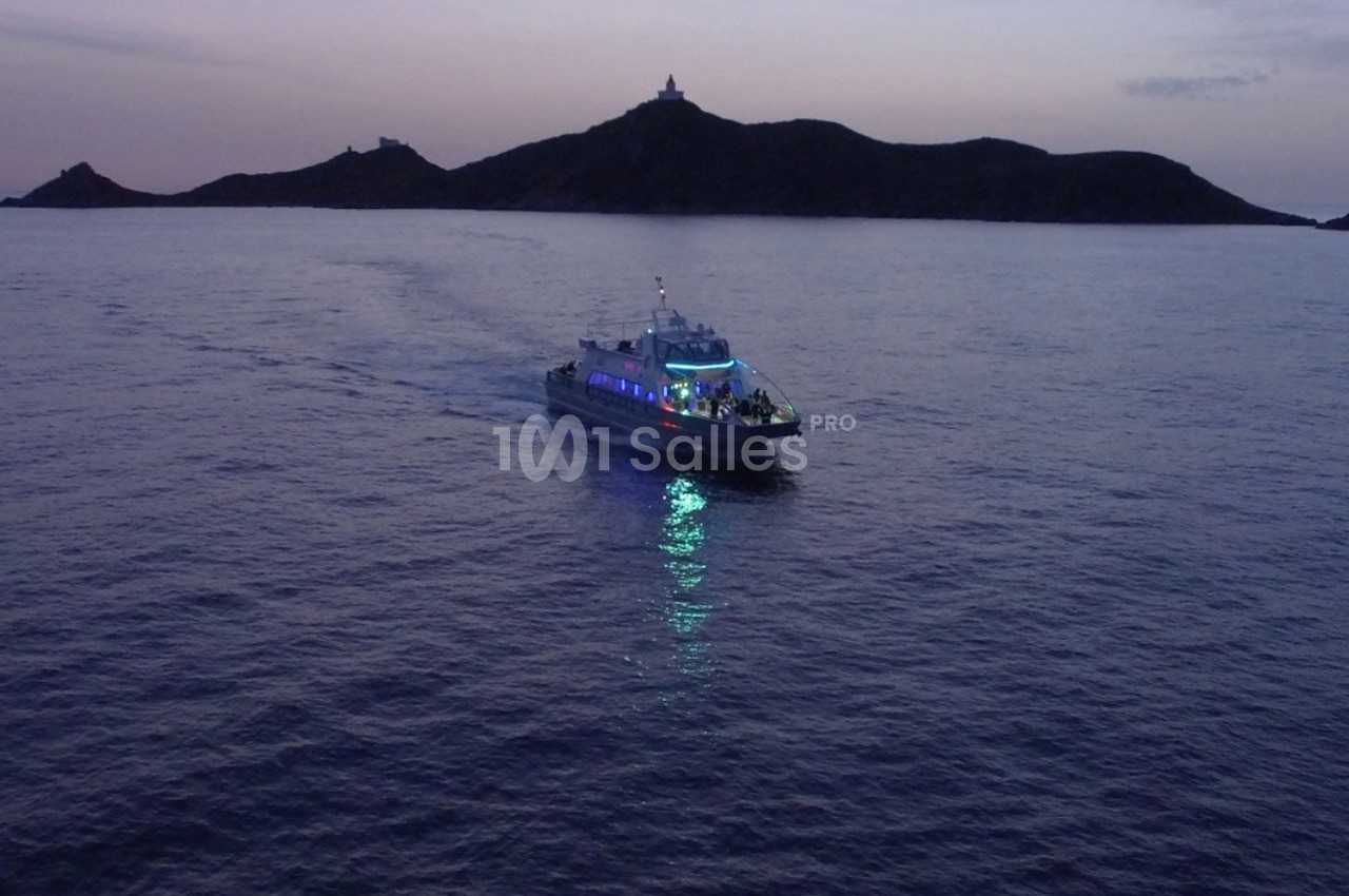 Un bateau éclairé navigue de nuit sur une mer calme, avec une île sombre en arrière-plan.