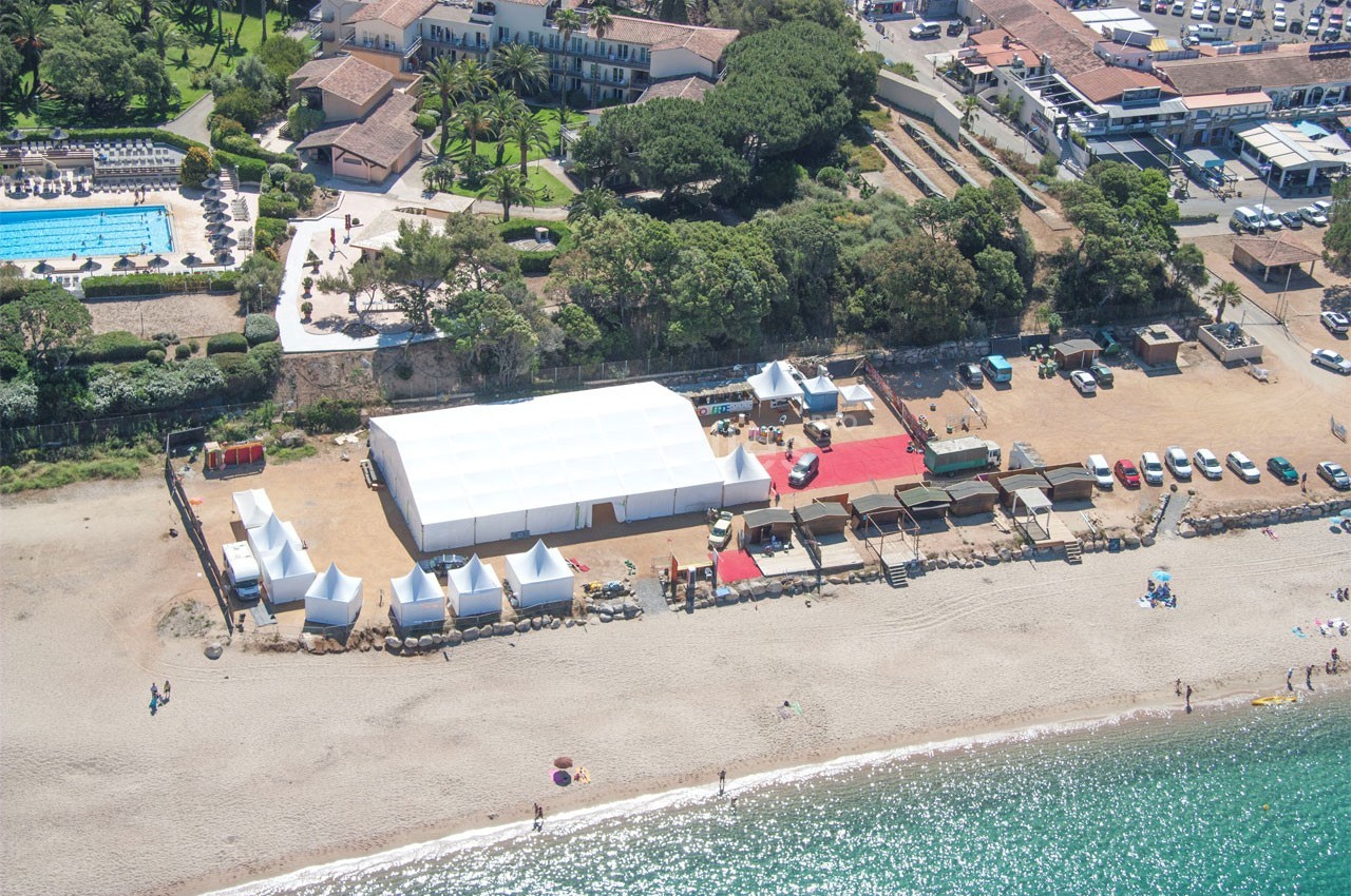 Vue aérienne d'une plage avec une grande structure blanche, des tentes, des voitures et des bâtiments en arrière-plan.