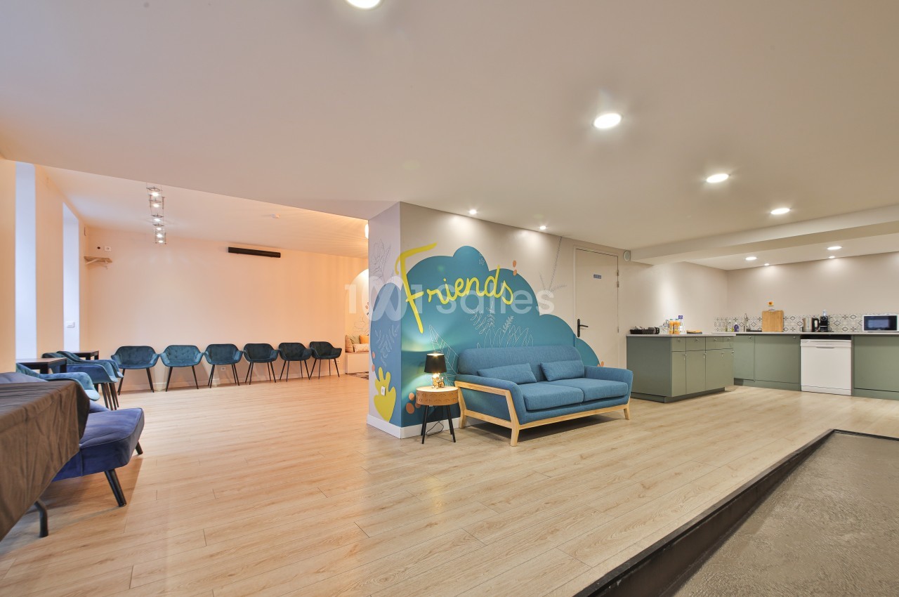 Salle lumineuse avec coin salon, kitchenette équipée et rangée de chaises alignées contre un mur clair.