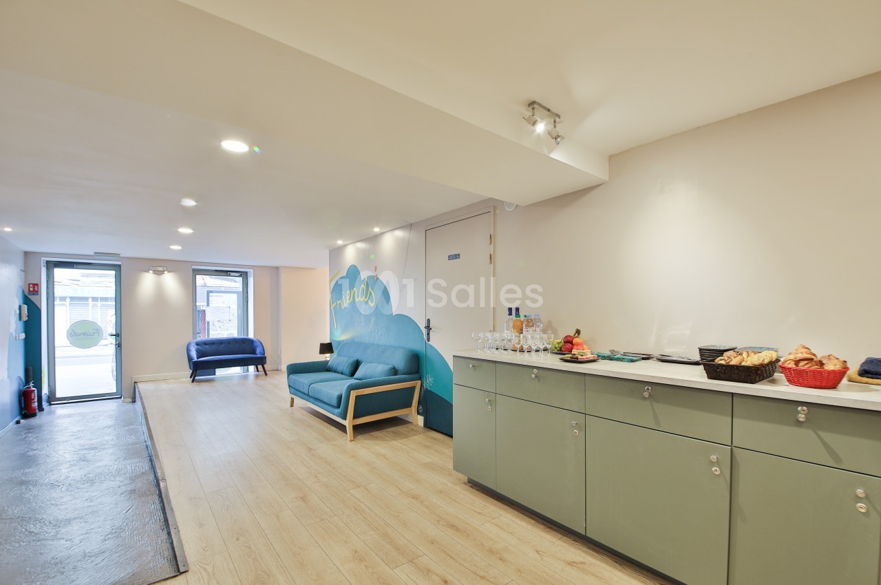 Salle lumineuse avec canapé bleu, mur décoré, buffet avec boissons et paniers de viennoiseries sur un meuble vert.