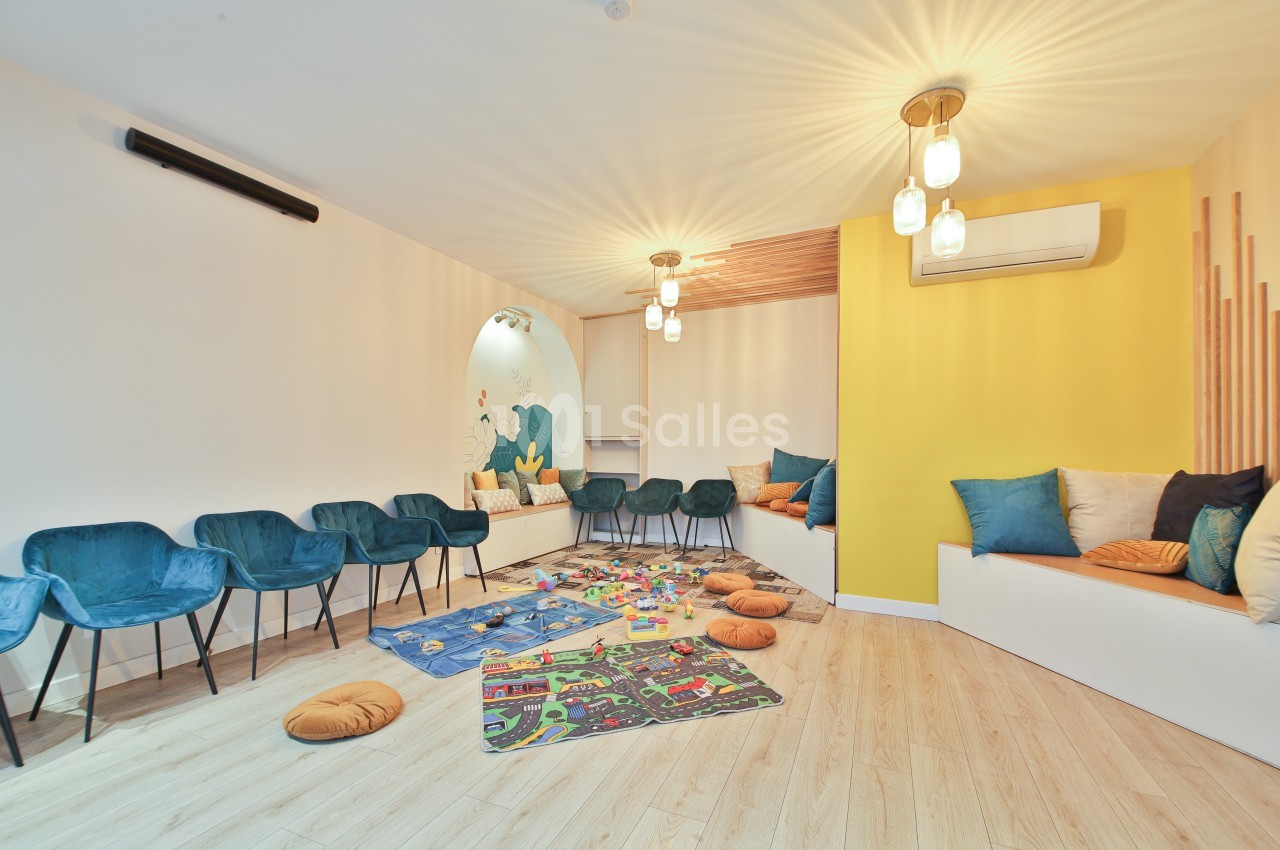 Salle lumineuse avec des sièges bleus, coussins colorés, tapis de jeu pour enfants et mur jaune décoratif.