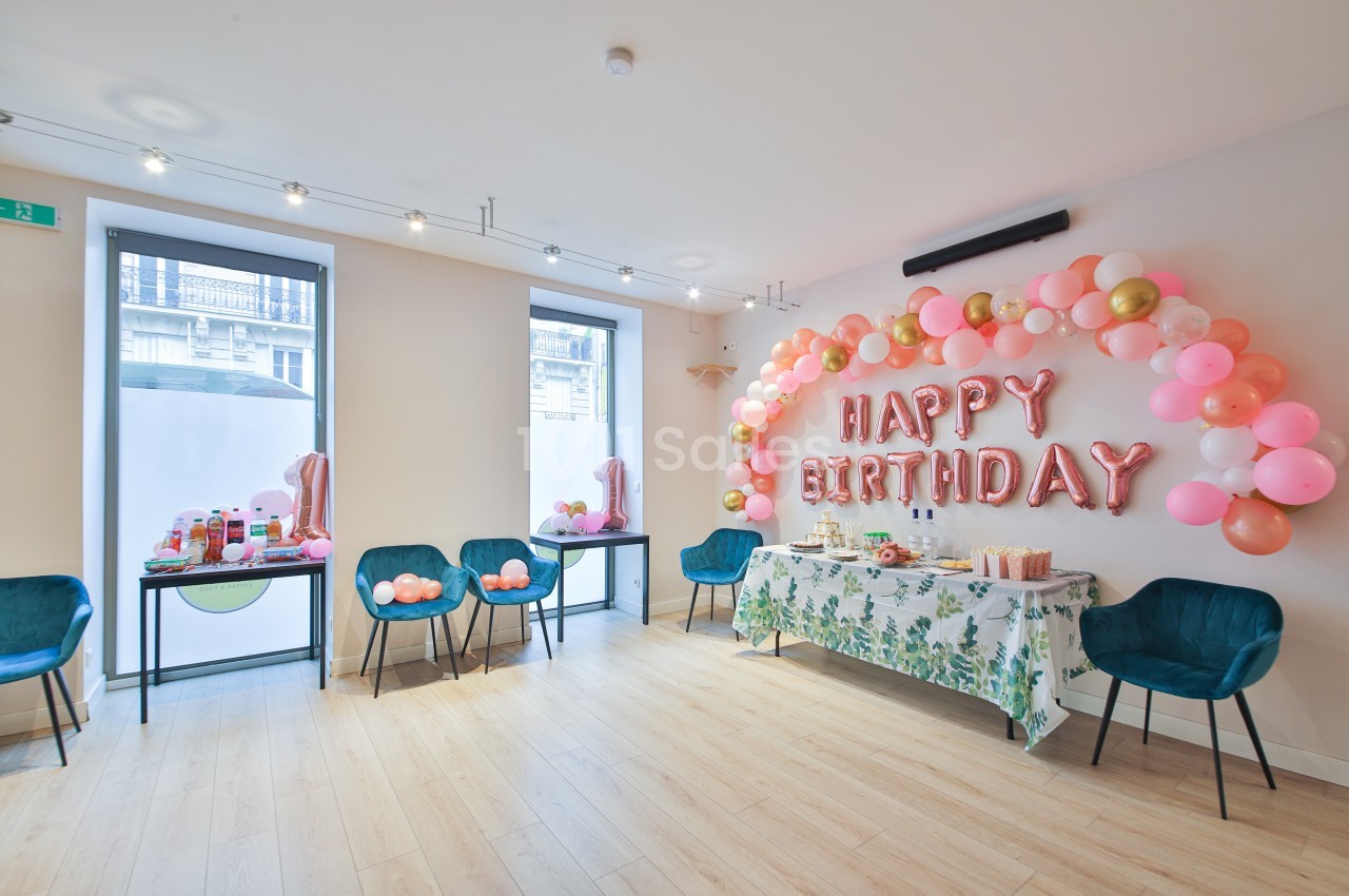 Salle décorée pour un anniversaire avec ballons roses et dorés, table garnie et chaises bleues.