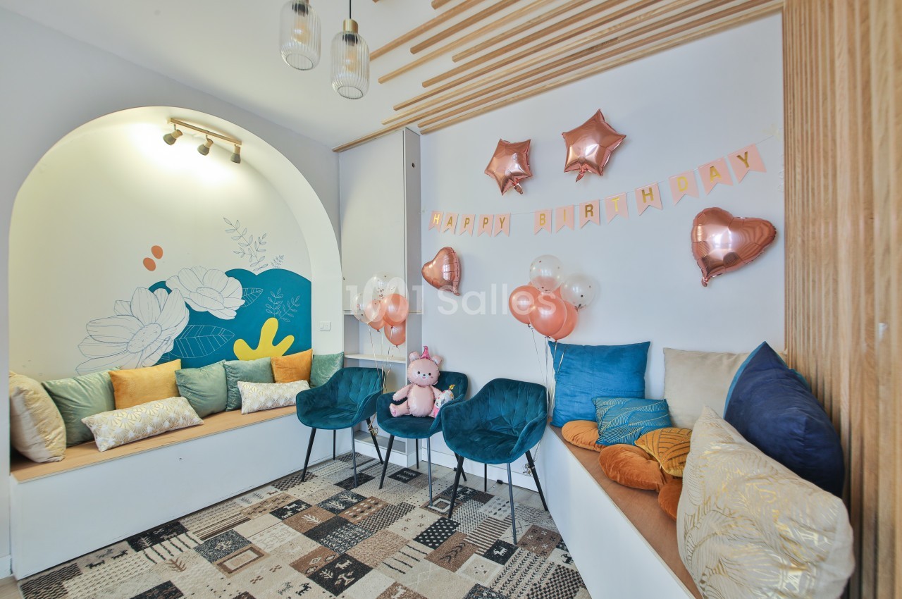 Salle décorée pour un anniversaire avec ballons roses, guirlande ’Happy Birthday’ et coussins colorés sur des banquettes.