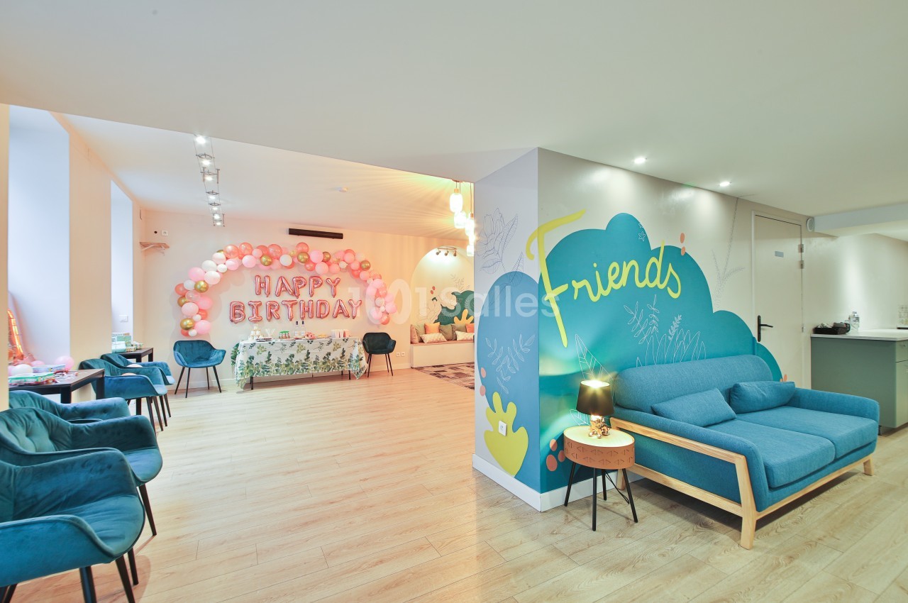 Salle lumineuse décorée pour un anniversaire, avec ballons, inscription ’Happy Birthday’ et coin salon coloré.