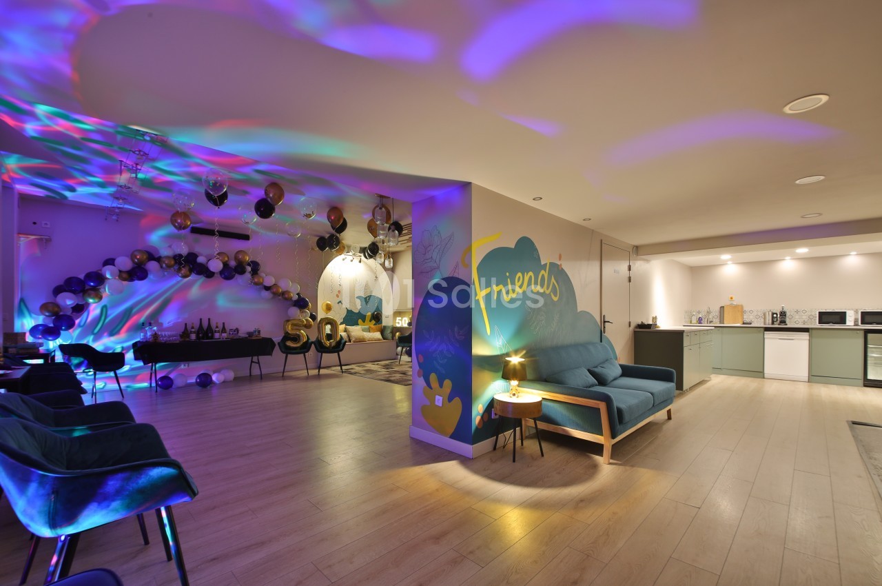 Salle décorée pour une fête avec ballons, éclairages colorés, coin salon et cuisine ouverte en arrière-plan.