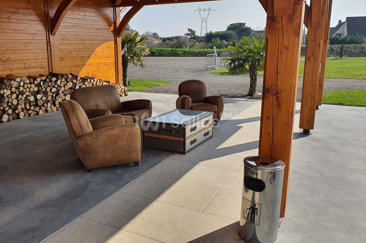 Salon extérieur avec fauteuils en cuir, table basse en métal et bois, sous une pergola en bois près d'un espace vert.